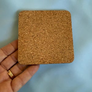 May include: A square cork coaster with rounded corners. The coaster is a light brown color and has a textured surface. The coaster is held by a hand with a gold ring.