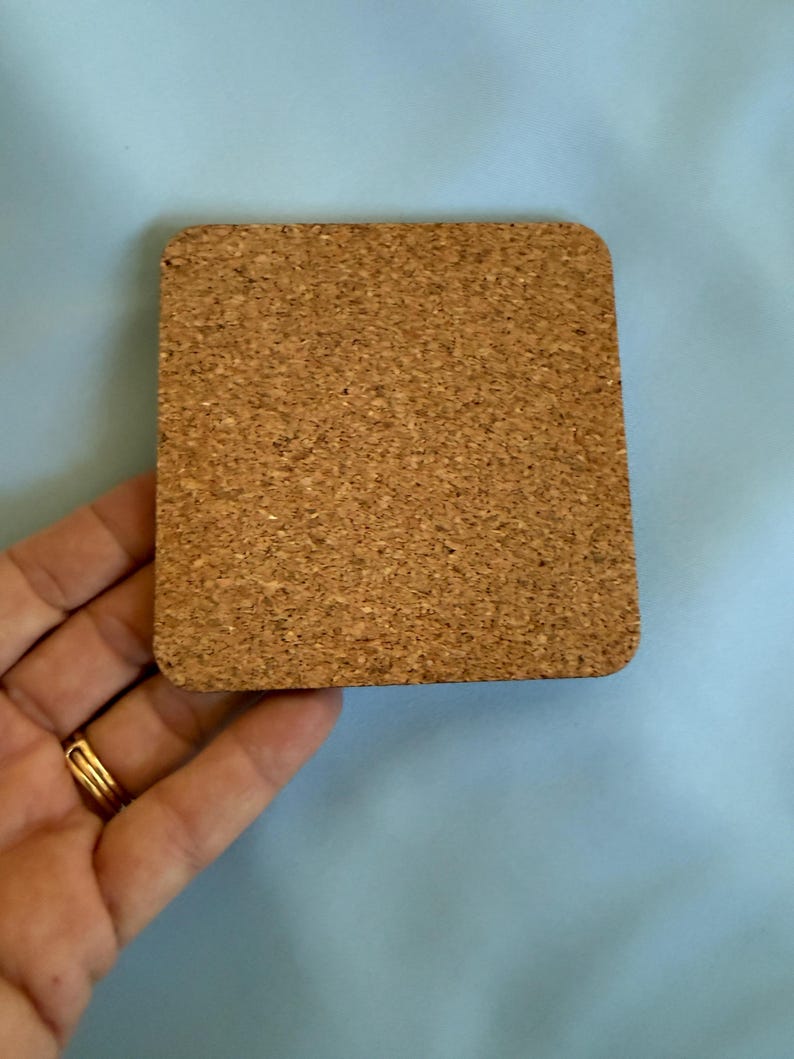 May include: A square cork coaster with rounded corners. The coaster is a light brown color and has a textured surface. The coaster is held by a hand with a gold ring.