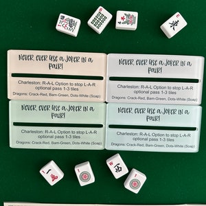 May include: Four white tiles with black Chinese characters and red dots. The tiles are arranged in a square shape. The tiles are on a green felt surface. The tiles are surrounded by four white cards with black text that reads "Never, ever use a joker in a pair!" and "Charleston: R-A-L Option to stop L-A-R optional pass 1-3 tiles Dragons: Crack-Red, Bam-Green, Dots-White (Soap)".