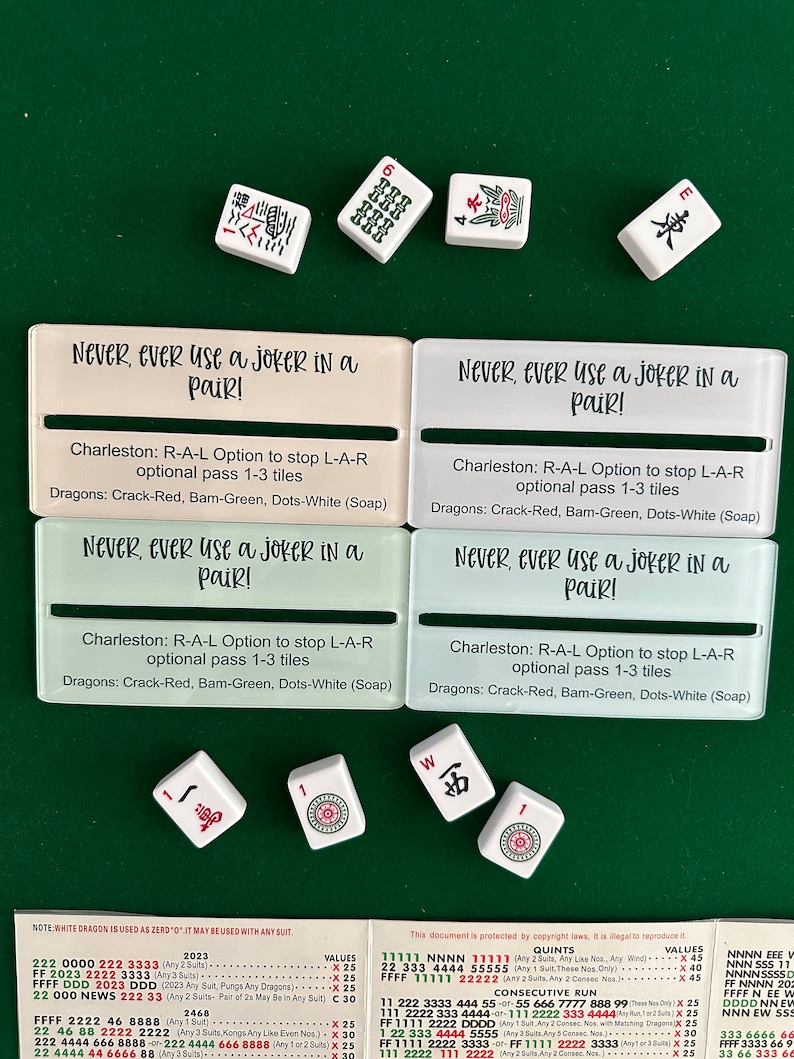 May include: Four white tiles with black Chinese characters and red dots. The tiles are arranged in a square shape. The tiles are on a green felt surface. The tiles are surrounded by four white cards with black text that reads "Never, ever use a joker in a pair!" and "Charleston: R-A-L Option to stop L-A-R optional pass 1-3 tiles Dragons: Crack-Red, Bam-Green, Dots-White (Soap)".