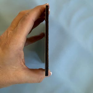 May include: A thin, rectangular wooden item with a dark brown edge and a lighter brown surface. The item is held between fingers against a light blue background. The item's thickness is visible.