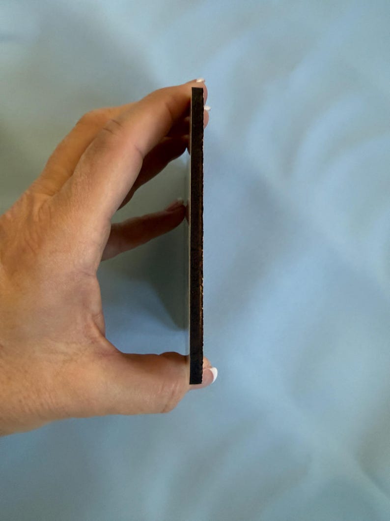 May include: A thin, rectangular wooden item with a dark brown edge and a lighter brown surface. The item is held between fingers against a light blue background. The item's thickness is visible.