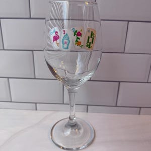 Pode incluir: Uma ta&ccedil;a de vinho de vidro transparente com um gr&aacute;fico decorativo de quatro pe&ccedil;as de mahjong dispostas em fila. As pe&ccedil;as exibem um flamingo cor-de-rosa, um biber&atilde;o azul com o n&uacute;mero 3, uma flor vermelha e caules de bambu verdes com o n&uacute;mero 4.