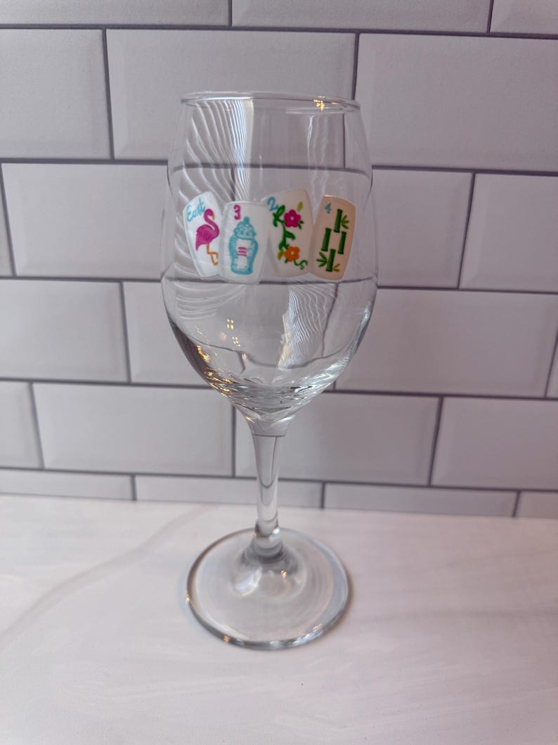 Pode incluir: Uma ta&ccedil;a de vinho de vidro transparente com um gr&aacute;fico decorativo de quatro pe&ccedil;as de mahjong dispostas em fila. As pe&ccedil;as exibem um flamingo cor-de-rosa, um biber&atilde;o azul com o n&uacute;mero 3, uma flor vermelha e caules de bambu verdes com o n&uacute;mero 4.