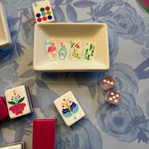 Może przedstawiać: Kolekcja płytek i kości do gry w Mahjong ułożonych na powierzchni z kwiatowym wzorem. Płytki mają r&oacute;żne kolorowe wzory, w tym kwiaty i cyfry. Mała biała miseczka zawiera dekoracyjną sztukę zdobienia paznokci.