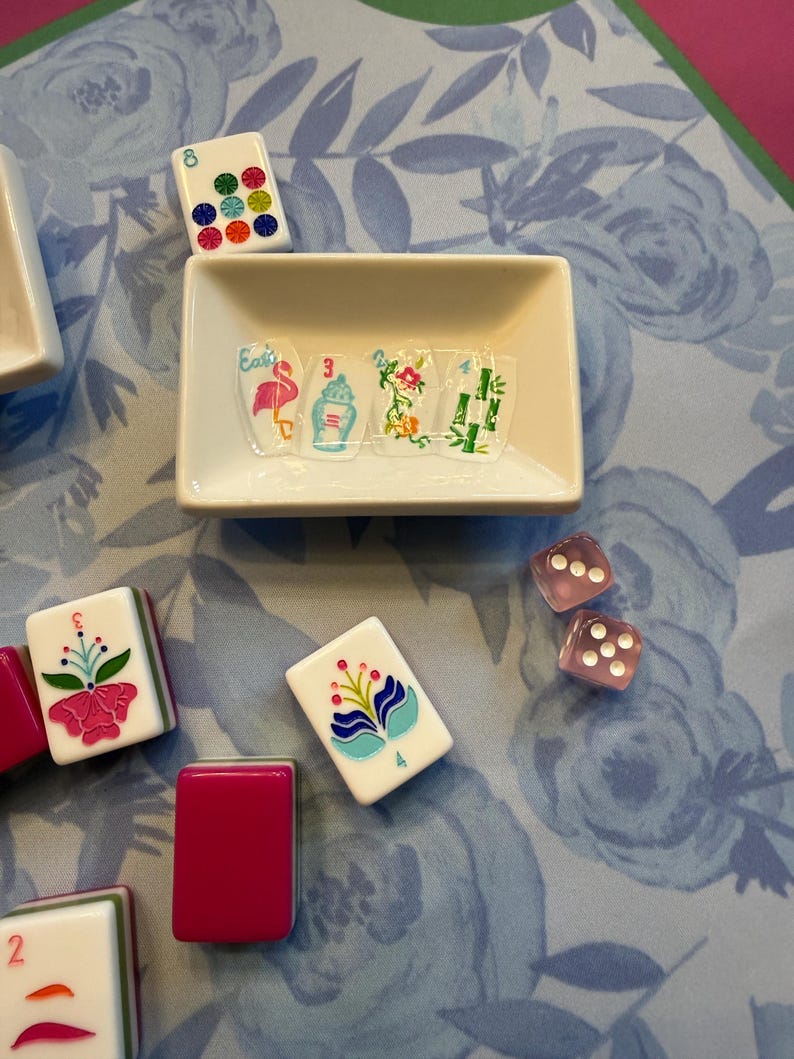 Może przedstawiać: Kolekcja płytek i kości do gry w Mahjong ułożonych na powierzchni z kwiatowym wzorem. Płytki mają r&oacute;żne kolorowe wzory, w tym kwiaty i cyfry. Mała biała miseczka zawiera dekoracyjną sztukę zdobienia paznokci.