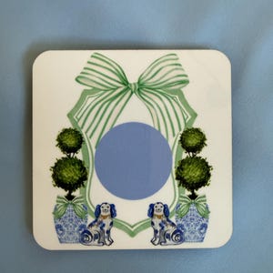 May include: Square white coaster with a blue and white design. Features a green bow, topiary trees, and two blue and white dog figurines. A blue circle is in the center of the design.