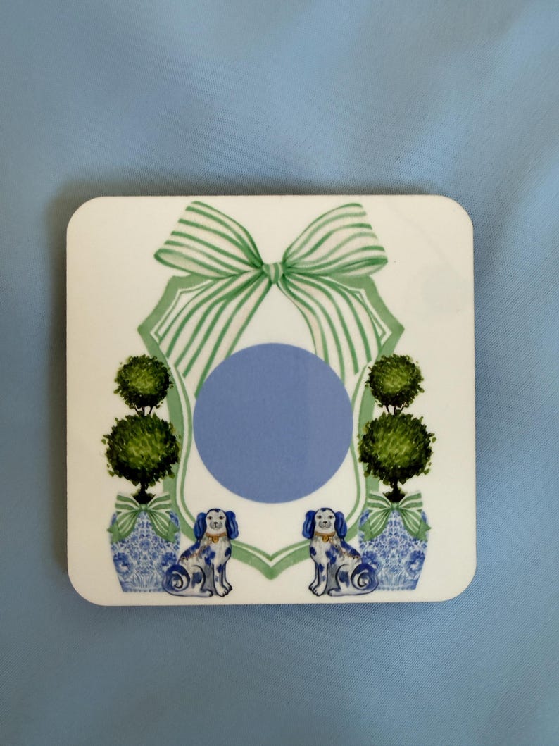 May include: Square white coaster with a blue and white design. Features a green bow, topiary trees, and two blue and white dog figurines. A blue circle is in the center of the design.