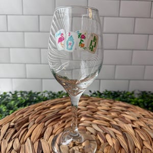 Pode incluir: Uma ta&ccedil;a de vinho de vidro transparente com quatro autocolantes de pe&ccedil;as de mahjong na parte frontal. Os desenhos incluem um flamingo cor-de-rosa, um n&uacute;mero tr&ecirc;s azul, um motivo floral e um s&iacute;mbolo de bambu verde, alinhados na horizontal.