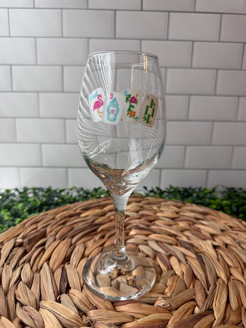 Pode incluir: Uma ta&ccedil;a de vinho de vidro transparente com quatro autocolantes de pe&ccedil;as de mahjong na parte frontal. Os desenhos incluem um flamingo cor-de-rosa, um n&uacute;mero tr&ecirc;s azul, um motivo floral e um s&iacute;mbolo de bambu verde, alinhados na horizontal.