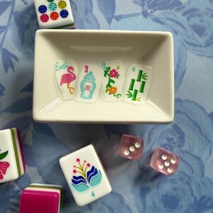 Może przedstawiać: Zestaw płytek do gry w Mahjong i r&oacute;żowych kostek do gry ułożonych na niebieskiej powierzchni w kwiatowy wz&oacute;r. Płytki mają kolorowe wzory, w tym flaminga, kwiaty i bambus. Biała prostokątna miska zawiera kilka płytek.