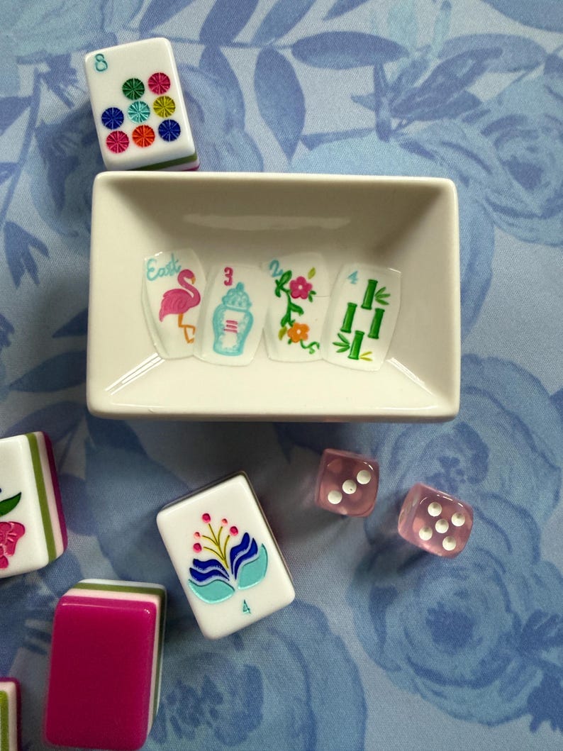 Może przedstawiać: Zestaw płytek do gry w Mahjong i r&oacute;żowych kostek do gry ułożonych na niebieskiej powierzchni w kwiatowy wz&oacute;r. Płytki mają kolorowe wzory, w tym flaminga, kwiaty i bambus. Biała prostokątna miska zawiera kilka płytek.