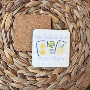 May include: A square cork coaster with a white and light yellow checkered surface featuring a graphic of three mahjong tiles, a lemon tree in a blue and white pot, and a glass of lemonade, with the text When Life gives you lemons, Play Mahjong.