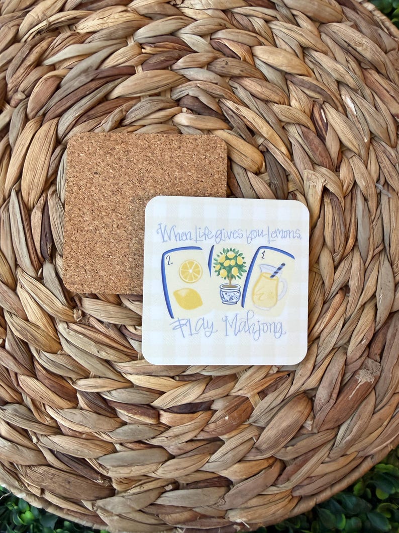 May include: A square cork coaster with a white and light yellow checkered surface featuring a graphic of three mahjong tiles, a lemon tree in a blue and white pot, and a glass of lemonade, with the text When Life gives you lemons, Play Mahjong.