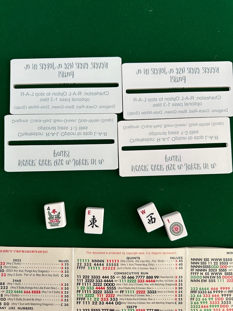 May include: A close-up of a Mahjong set with four tiles on a green felt surface. The tiles are white with black characters and red, green, and white dots. The tiles are labeled "4", "E", "W", and "1". The tiles are arranged in a row. The image also includes a chart with Mahjong hand values and rules.