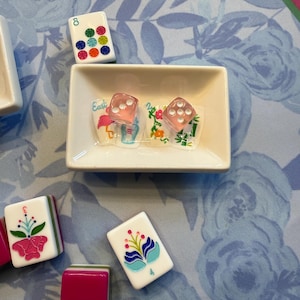 Może przedstawiać: Zestaw płytek i kości do gry w Mahjong ułożonych na powierzchni z kwiatowym wzorem. Płytki mają kolorowe kwiatowe wzory i liczby. Dwie r&oacute;żowe kości do gry znajdują się w małej białej miseczce ze słowem "Easter" na naklejce. Tło to niebieski kwiatowy wz&oacute;r.