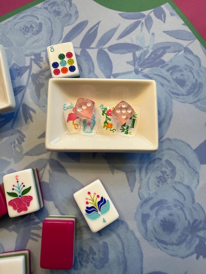Może przedstawiać: Zestaw płytek i kości do gry w Mahjong ułożonych na powierzchni z kwiatowym wzorem. Płytki mają kolorowe kwiatowe wzory i liczby. Dwie r&oacute;żowe kości do gry znajdują się w małej białej miseczce ze słowem "Easter" na naklejce. Tło to niebieski kwiatowy wz&oacute;r.
