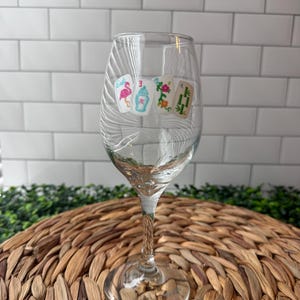 Pode incluir: Uma ta&ccedil;a de vinho de vidro transparente com um design impresso de quatro pe&ccedil;as de mahjong. As pe&ccedil;as exibem um flamingo cor-de-rosa, uma coqueteleira azul e branca, um padr&atilde;o floral colorido e caules de bambu verdes, com haste fina.