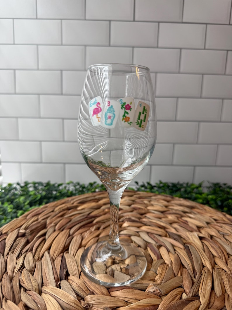 Pode incluir: Uma ta&ccedil;a de vinho de vidro transparente com um design impresso de quatro pe&ccedil;as de mahjong. As pe&ccedil;as exibem um flamingo cor-de-rosa, uma coqueteleira azul e branca, um padr&atilde;o floral colorido e caules de bambu verdes, com haste fina.