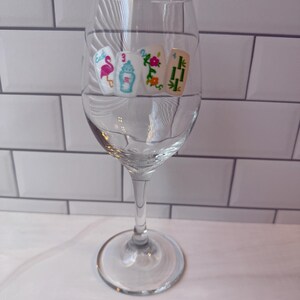 Pode incluir: Uma ta&ccedil;a de vinho de vidro transparente com quatro decalques de pe&ccedil;as de Mahjong dispostos numa fila horizontal. As pe&ccedil;as exibem um flamingo cor-de-rosa, uma urna azul, um padr&atilde;o floral e caules de bambu verdes, com n&uacute;meros e texto colorido.
