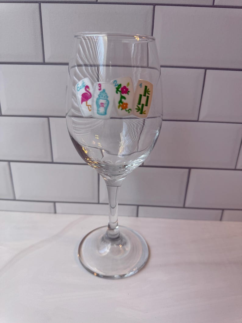 Pode incluir: Uma ta&ccedil;a de vinho de vidro transparente com quatro decalques de pe&ccedil;as de Mahjong dispostos numa fila horizontal. As pe&ccedil;as exibem um flamingo cor-de-rosa, uma urna azul, um padr&atilde;o floral e caules de bambu verdes, com n&uacute;meros e texto colorido.