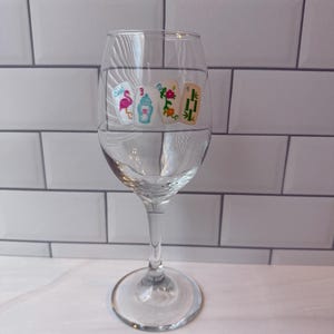 Pode incluir: Uma ta&ccedil;a de vinho de vidro transparente com um decalque decorativo de quatro pe&ccedil;as de mahjong. As pe&ccedil;as exibem um flamingo cor-de-rosa com a palavra East, um n&uacute;mero tr&ecirc;s azul, uma flor vermelha e um desenho de bambu verde com o n&uacute;mero quatro.