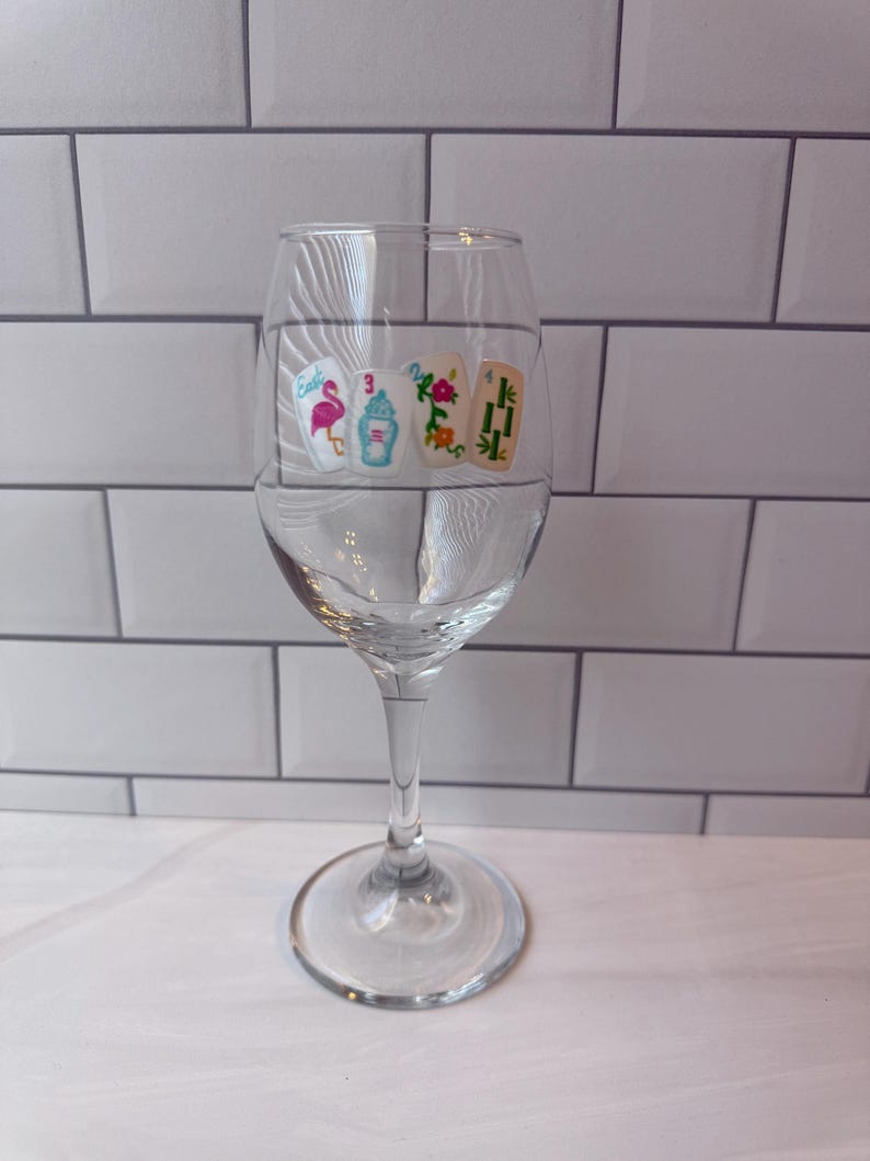 Pode incluir: Uma ta&ccedil;a de vinho de vidro transparente com um decalque decorativo de quatro pe&ccedil;as de mahjong. As pe&ccedil;as exibem um flamingo cor-de-rosa com a palavra East, um n&uacute;mero tr&ecirc;s azul, uma flor vermelha e um desenho de bambu verde com o n&uacute;mero quatro.