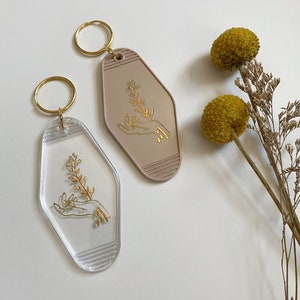 May include: Two keychains with a gold outline of a hand holding a flower sprig. One keychain is clear plastic and the other is a light brown plastic.
