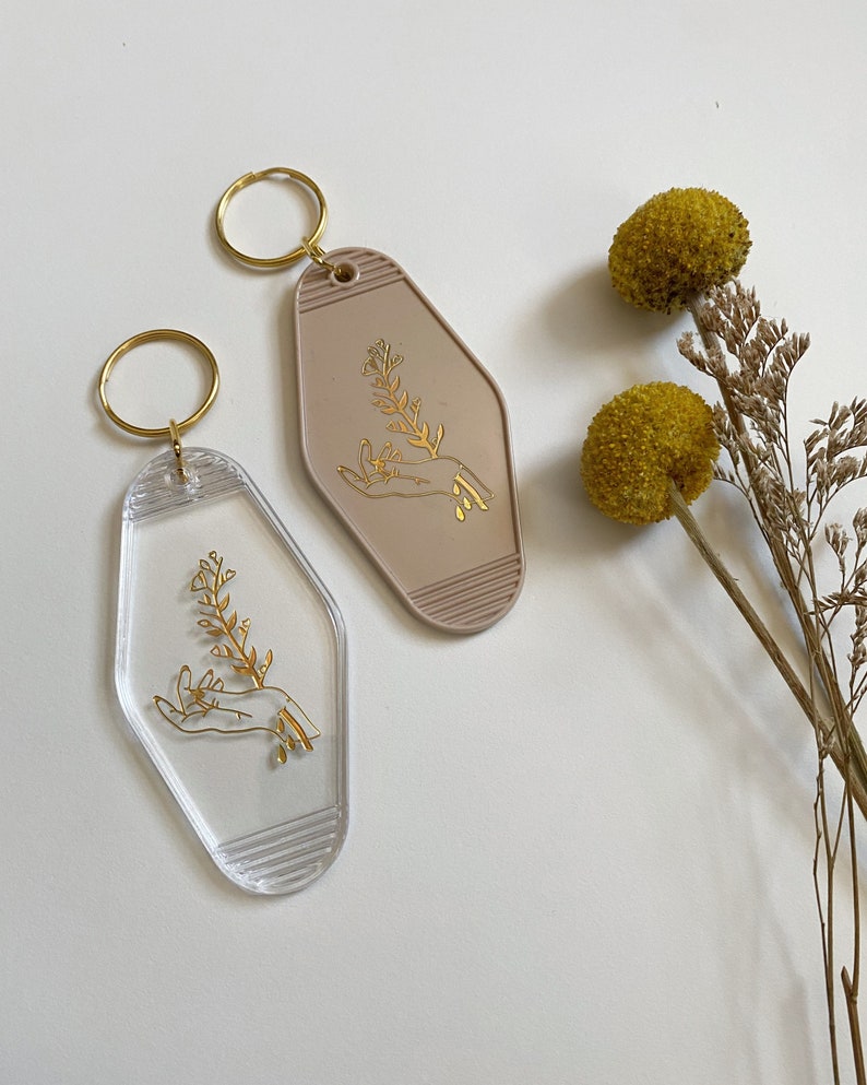 May include: Two keychains with a gold outline of a hand holding a flower sprig. One keychain is clear plastic and the other is a light brown plastic.