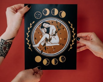 May include: Black art print depicting a woman with moon phases and stars. The illustration uses gold and brown tones against a black backdrop. A decorative laurel wreath encircles the central image.
