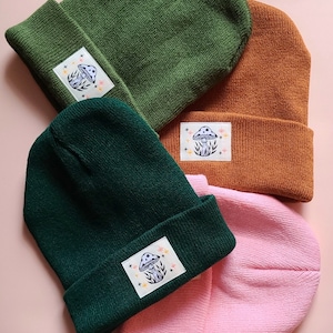 Könnte beinhalten: Drei Strickmützen in verschiedenen Farben: Grün, Dunkelgrün und Rosa. Jede Mütze hat einen Pilz-förmigen Aufnäher mit einem weißen Pilz und bunten Details.