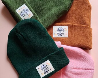 May include: Three knitted beanies in different colors: green, dark green, and pink. Each beanie has a mushroom-shaped patch with a white mushroom and colorful details.