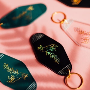 May include: Five keychains with gold foiled designs on a pink background. The keychains are shaped like hotel room keys and feature a hand holding a plant, a floral design, and a geometric pattern.
