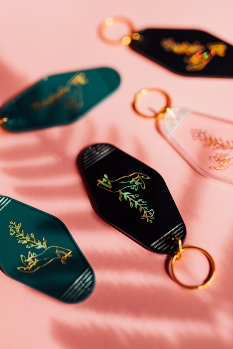 May include: Five keychains with gold foiled designs on a pink background. The keychains are shaped like hotel room keys and feature a hand holding a plant, a floral design, and a geometric pattern.