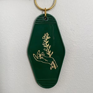 May include: A green motel key chain with a gold-colored design of a hand holding a flower.