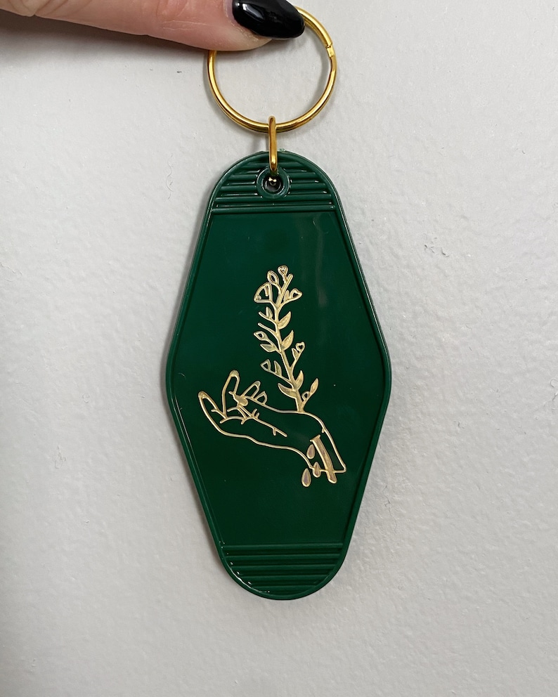 May include: A green motel key chain with a gold-colored design of a hand holding a flower.