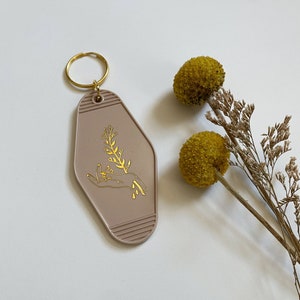 May include: A light brown motel key chain with a gold design of a hand holding a sprig of flowers. The keychain has a gold metal ring.