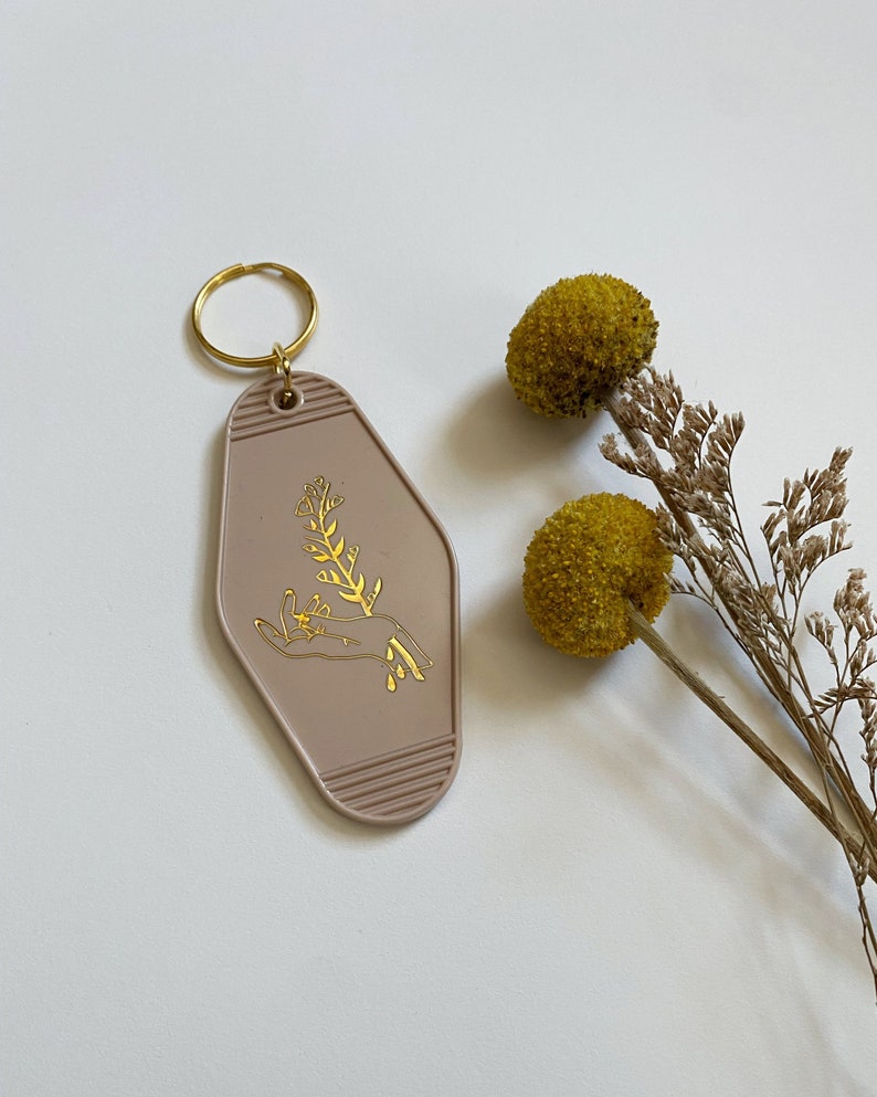 May include: A light brown motel key chain with a gold design of a hand holding a sprig of flowers. The keychain has a gold metal ring.