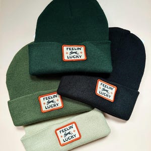 May include: Five knitted beanies in shades of green and black. Each beanie has a rectangular patch with the words "FEELIN' LUCKY" and a rabbit graphic. The colours include dark green, olive, and light green.