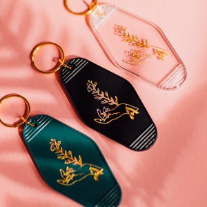 May include: Three keychains with a gold hand holding a sprig of flowers. The keychains are in three different colors: clear, black, and green.