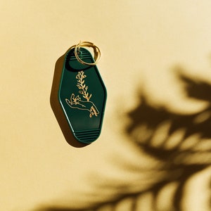 May include: A dark green motel key chain with a gold ring. The keychain features a gold outline of a hand holding a small plant.