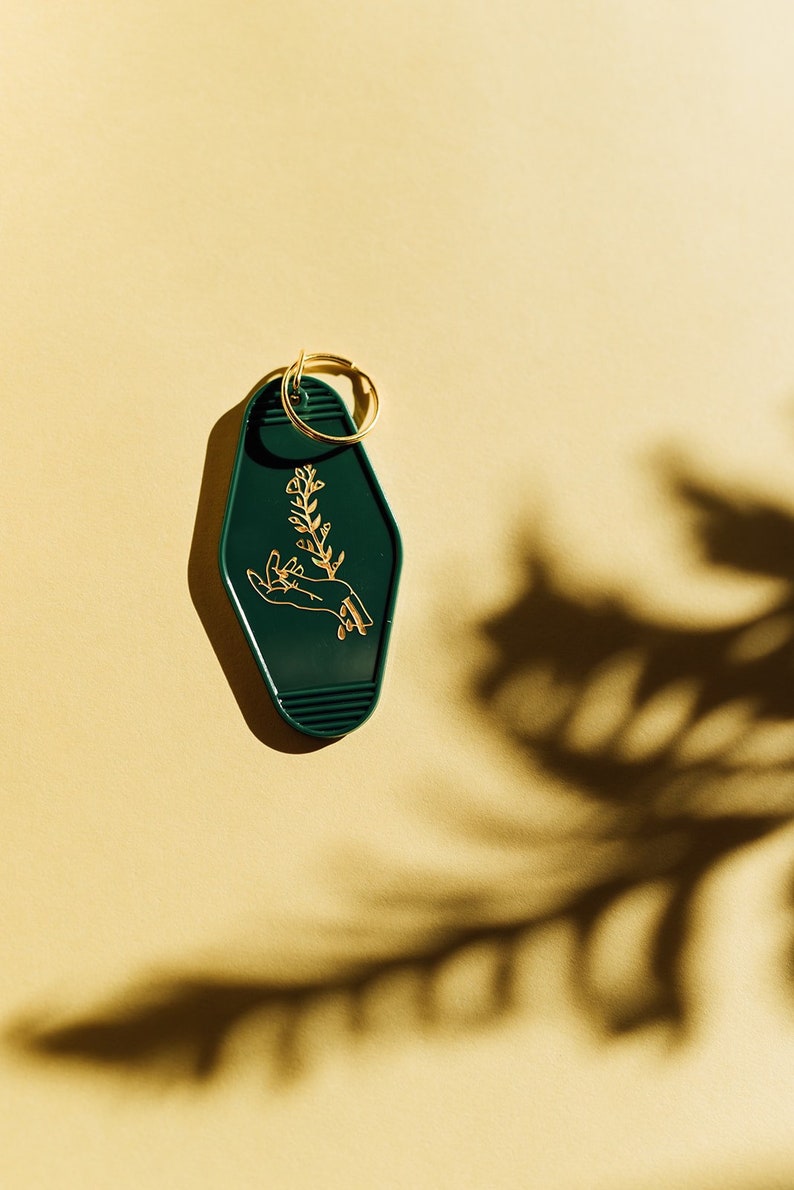 May include: A dark green motel key chain with a gold ring. The keychain features a gold outline of a hand holding a small plant.
