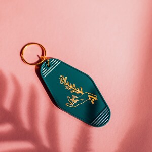 May include: A teal blue motel key tag with a gold outline and a gold design of a hand holding a plant.