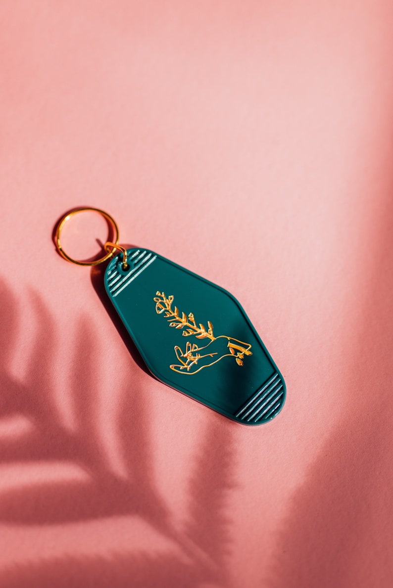 May include: A teal blue motel key tag with a gold outline and a gold design of a hand holding a plant.