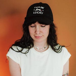 以下が含まれることがあります： 「FEELIN' LUCKY」の文字と白いウサギのグラフィックが入った黒い野球帽。白いTシャツを着た人がかぶっています。