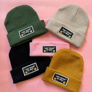 May include: A collection of beanies in various colors, including green, tan, pink, black, and mustard yellow. Each beanie has a rectangular patch with the text "JUST TRYING MY BEST" and a smiley face and frowny face.