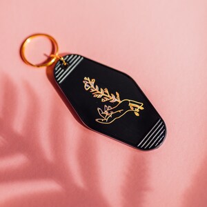 May include: Black motel key chain with a gold ring. The keychain features a gold outline of a hand holding a sprig of flowers.