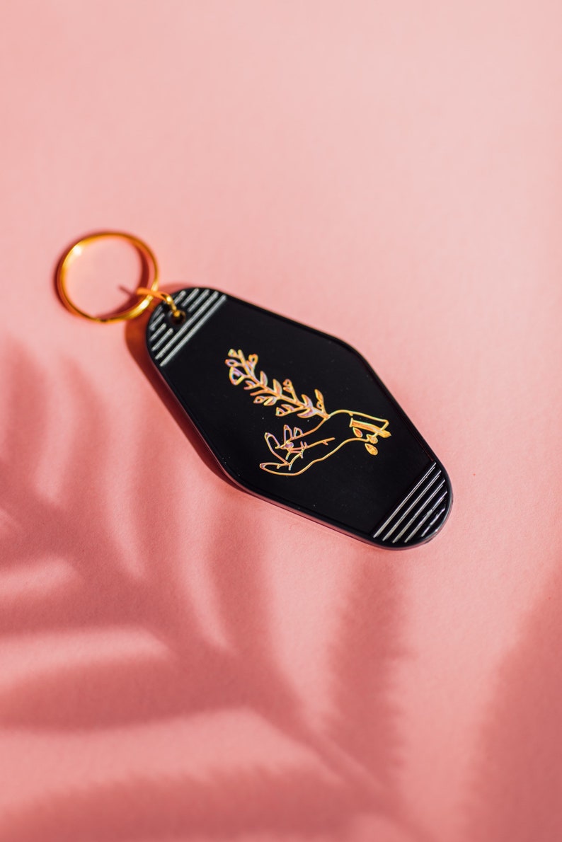 May include: Black motel key chain with a gold ring. The keychain features a gold outline of a hand holding a sprig of flowers.