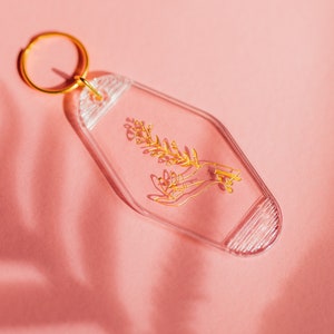 May include: A clear plastic keychain with a gold-colored design of a hand holding a sprig of flowers. The keychain has a gold-colored ring.