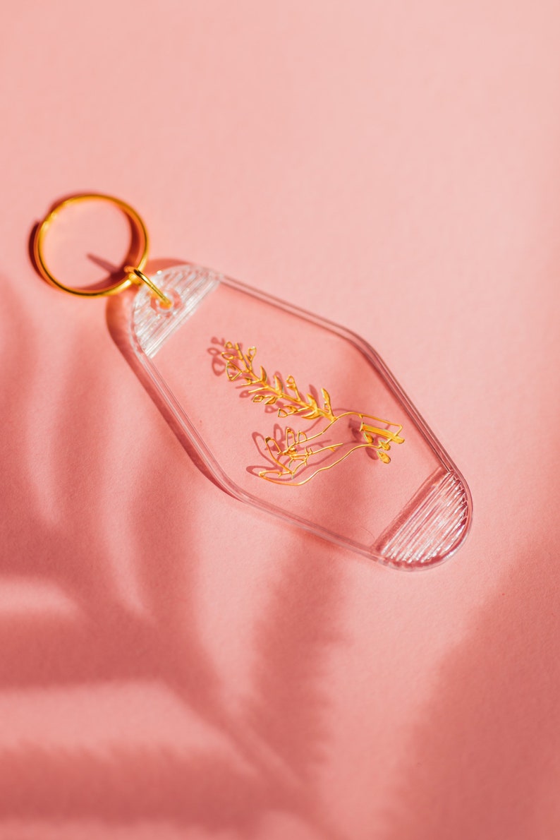 May include: A clear plastic keychain with a gold-colored design of a hand holding a sprig of flowers. The keychain has a gold-colored ring.